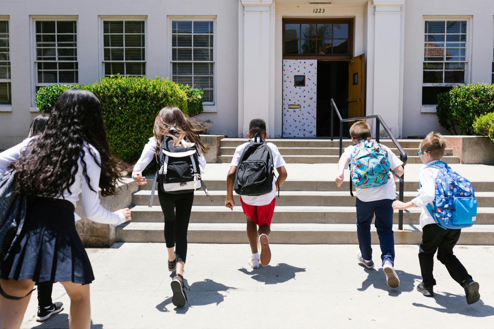 Five children with backpacks running up the front steps toward a school entrance.